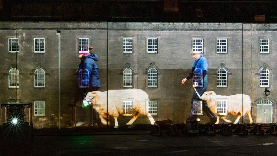 A projection on the walls of the barracks shows two women in blue clothing walking sheep