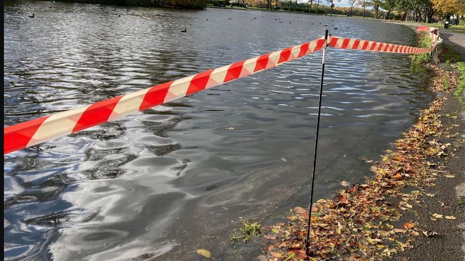 Lake at Pontefract Park with ducks on the water, a person walking a dog on the parth to the right, autumn leaves on the floor, red and white safety tape to cordon off the edge.