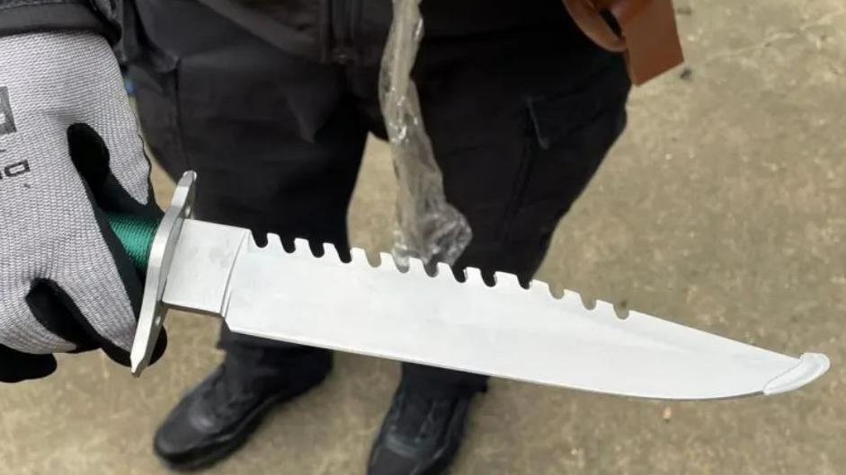 Person holding a large serrated knife while wearing grey gloves.