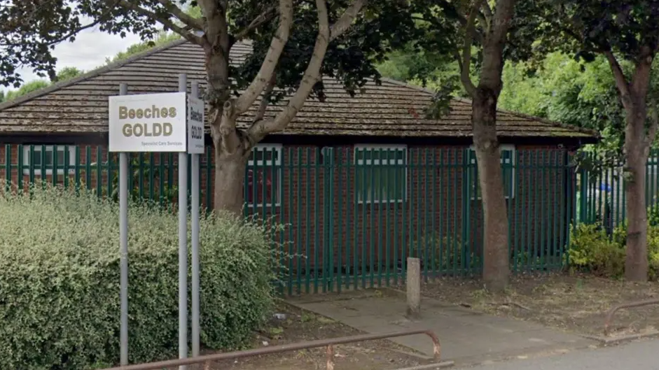 The sign for Beaches GOLDD centre. The building is surrounded by a green fence with a path leading up to it. Three trees can be seen in front of the centre and a small post. 