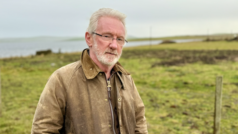 a man with grey hair, a beard and glasses is wearing a weathered-looking jacket and is standing in a field next to a stretch of water