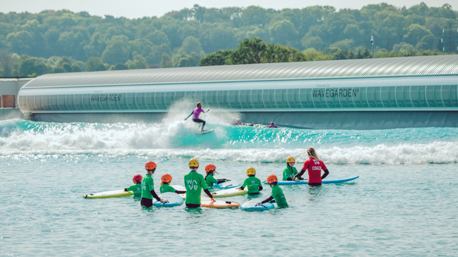 A group of children wearing helmets and green t-shirts stand in the water with a surfboard each, surrounding an instructor wearing a red t-shirt.