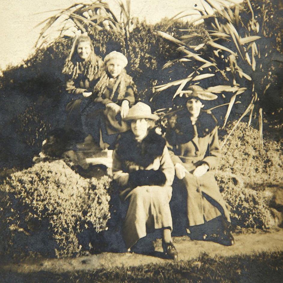 A black and white photo of four people on an outing sitting on what appears to be a hedge next to a conifer. Two older woman are wearing long coats and hats and two girls behind them are wearing scarves and coats.