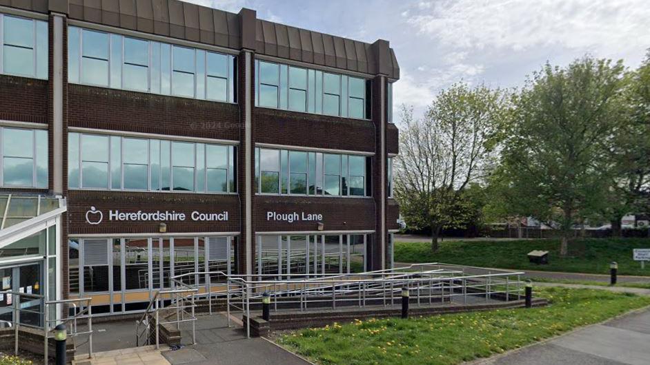 Herefordshire Council's offices in a modern building with large windows and a ramp with railings at the front. There is grass at the front of the building and an open space with trees to one side.