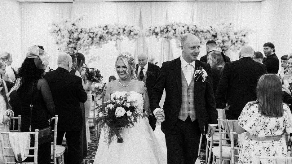 Lisa Marsay smiles in a wedding dress while holding her husband's hand and walking down the aisle at her wedding. 