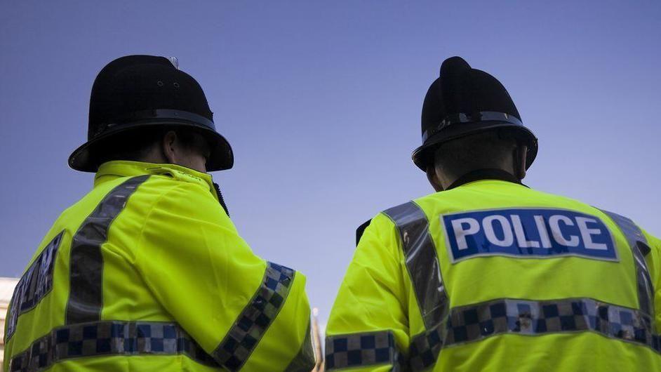 Two police officers facing away from the camera. They are wearing hi-vis police jackets with the word "police" written across the back and they are wearing black police helmets.