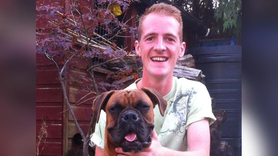 Sam Boulton wearing a green t shirt smiling with his hand on a brown dog, which has its tongue out.