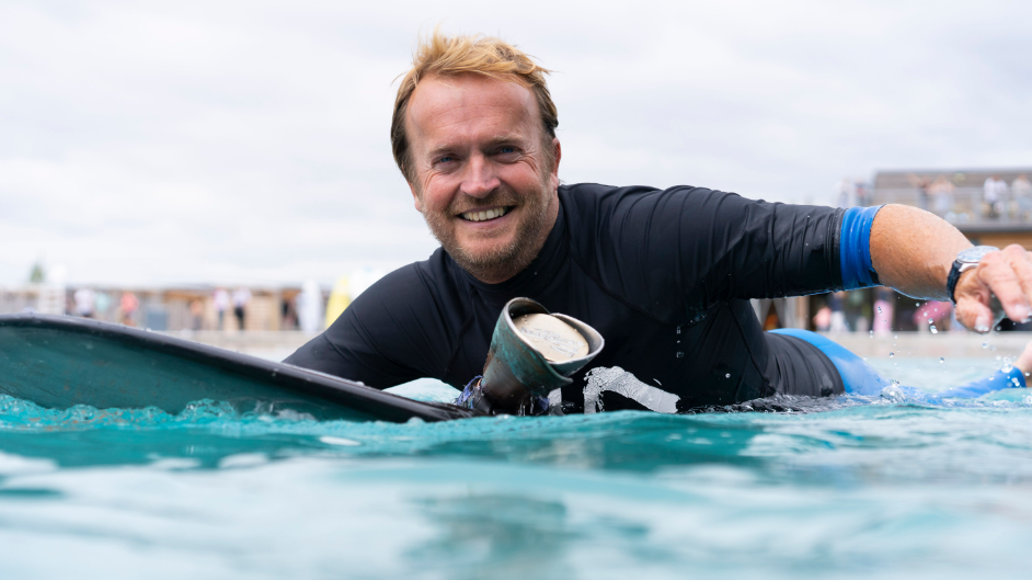 Nick Housfield smiles into the camera as he paddles out on his surfboard 