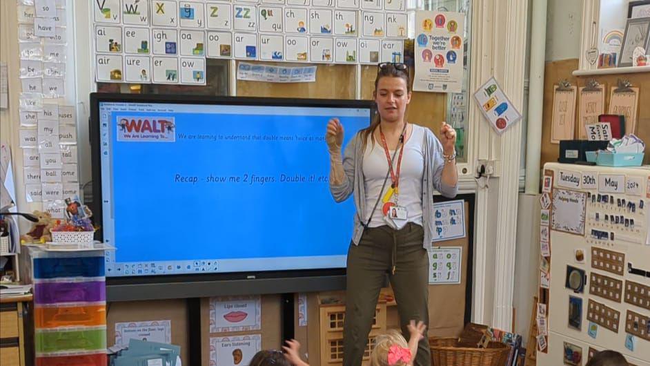 Amy stood at the front of a class in front of a board and teaching children