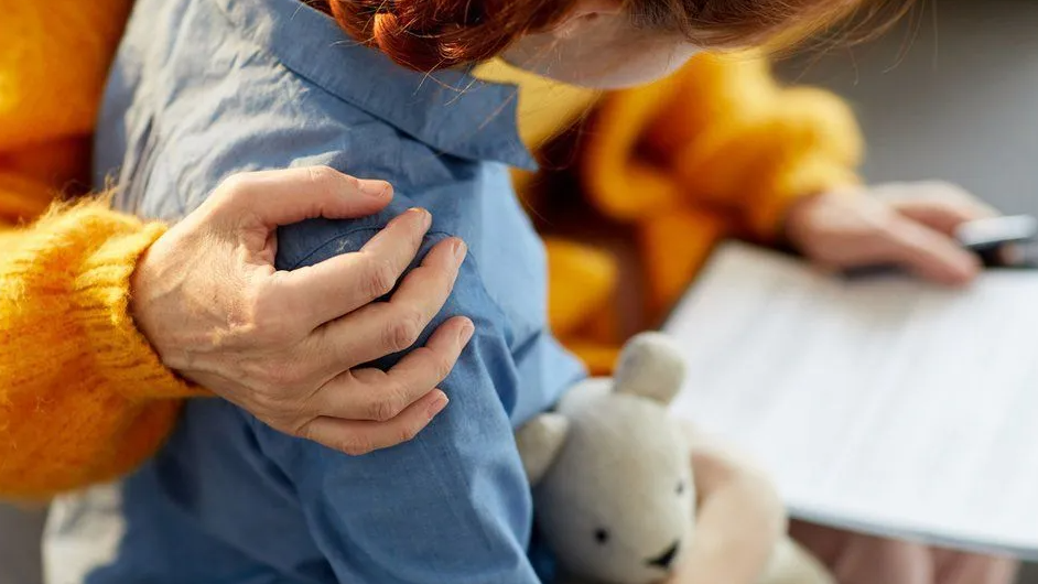 Child in a blue shirt, holding a teddy bear with a woman in an orange cardigan putting one arm around the child
