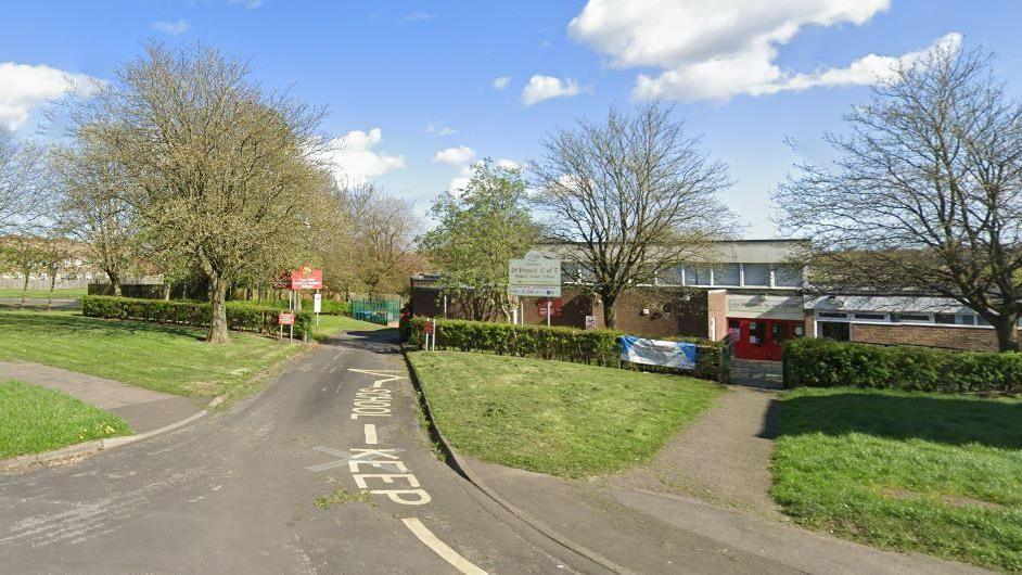 Entrance drive to the two schools. There is a "Keep Clear" sign painted on the road. The school buildings are surrounded by trees and grass.