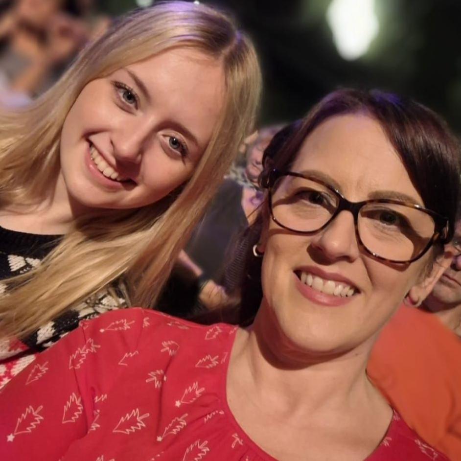 A selfie of Helen, a brunette woman with glasses, wearing a red dress, and Meg beside her, with long blonde hair. They are both smiling and looking at the camera.