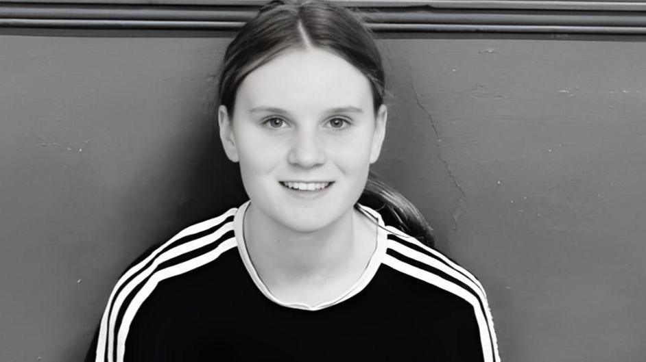 Holly Newton, 15, wearing a black top with white stripes on the shoulder. She is pictured smiling, with her hair pulled back in a ponytail 