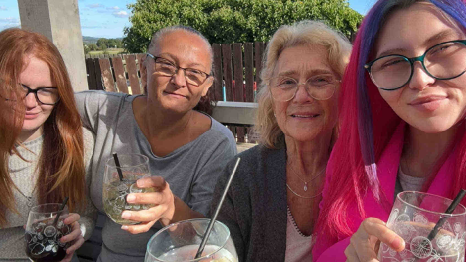 Four people sit outdoors at a table, each holding a drink in a clear glass with a straw. They are all dressed casually, with Jasmin having vibrant pink and purple hair. Behind them, there is a wooden fence and trees under a blue sky.