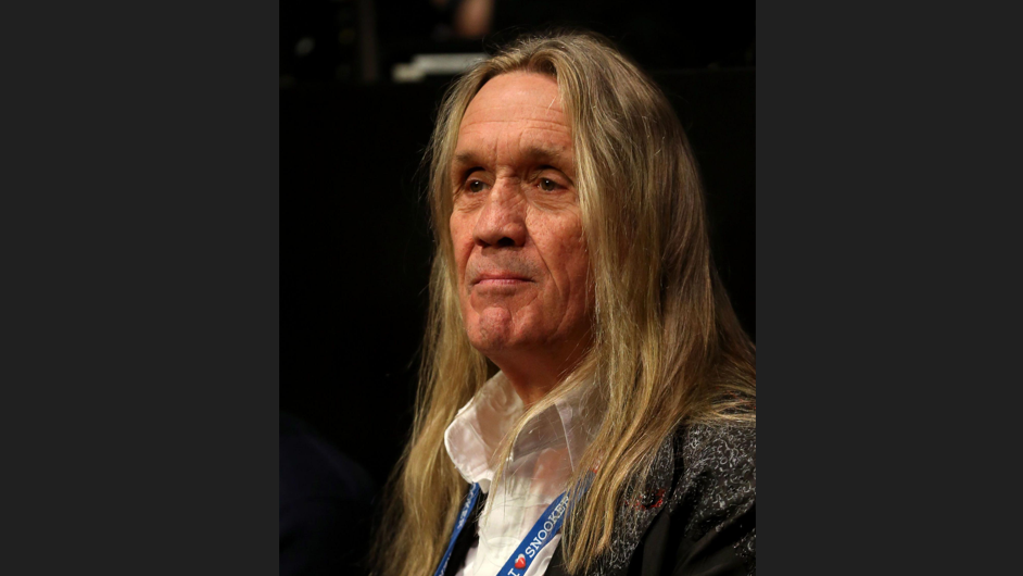 A head-and-shoulders picture of Nicko McBrain. He has long brown hair and is looking pensive. He is wearing an open-necked white shirt and a jacket.