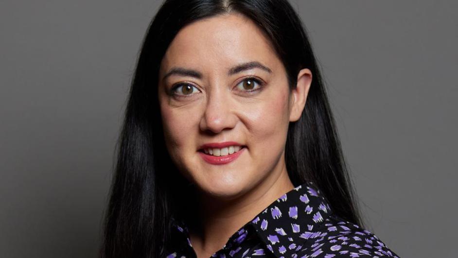 Sarah Owen MP looks directly at the camera. She has long dark hair, red lipstick and is wearing a black and mauve patterned blouse. Behind her is a plain grey backdrop.