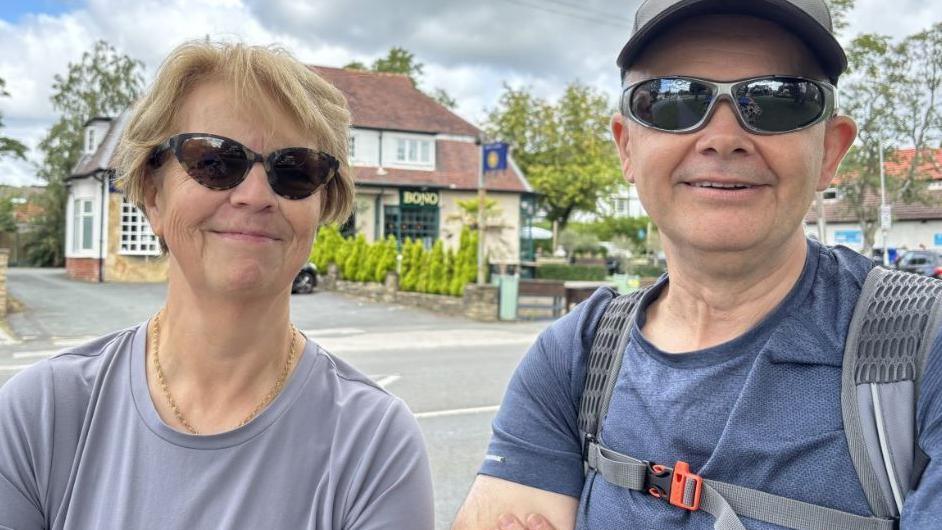 A man and woman are standing with a busy main road in the background. 
The woman has short blonde hair and is wearing sunglasses.
The man has a grey hat and sunglasses. He is wearing a blue t-shirt and carrying a rucksack. 