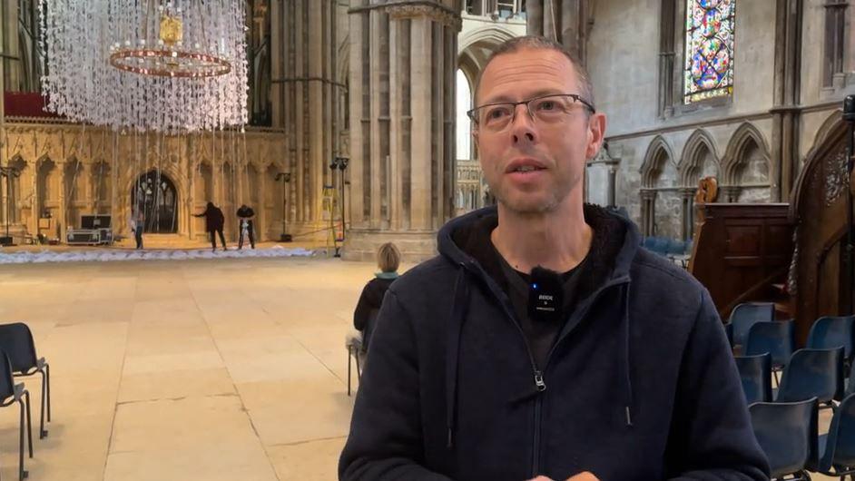 Artist Peter Walker in a black fleece and microphone on his t shirt with short hair and rectangular glasses looking at a reporter out of shot with the cathedral in the background and the white paper doves hanging from the nave in the background