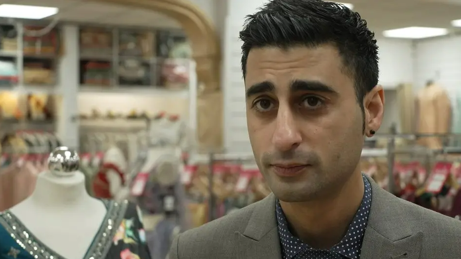 Karan Modha, wearing a grey suit jacket with a blue and white polka dot shirt underneath standing inside his saree shop. 