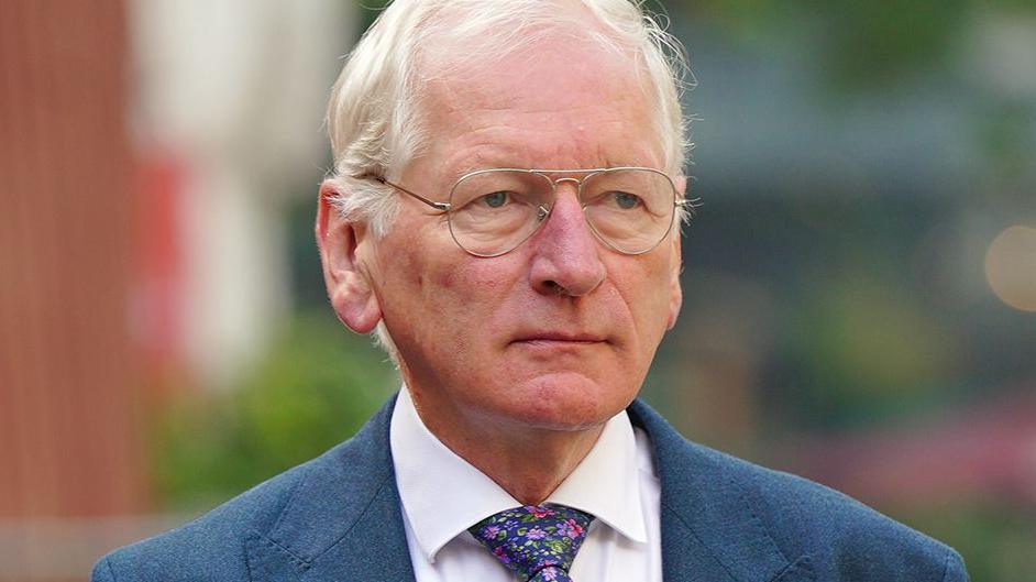 Dr Dewi Evans with grey hair wearing glasses a blue suit jacket, white shirt and blue, purple and pink flowery tie looking impassively