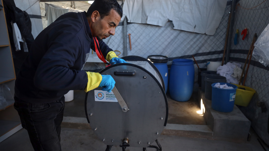 A member of the hospital team wearing navy blue clothes and blue sterile gloves  using the hand crank to make the washing machine work. The washing machine is like a big metal drum on its side with the Washing Machine Project written on it.