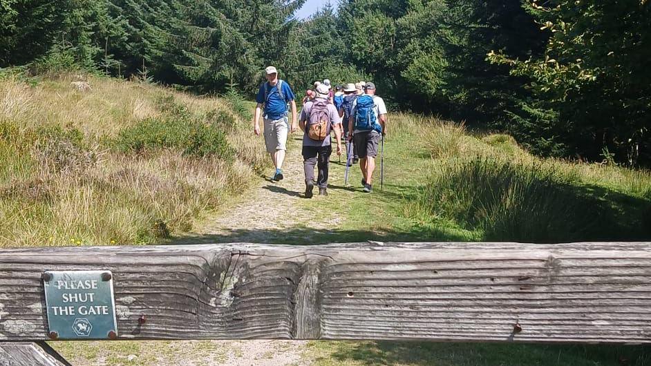 A group of ramblers walk past a man going in the opposite direction along a public footpath on Dartmoor. The man is wearing a grey baseball cap, blue t-shirt and beige shorts. A wooden gate is in the foreground of the photo with a small green sign on it saying "PLEASE SHUT THE GATE" with a Dartmoor National Park logo underneath the writing.