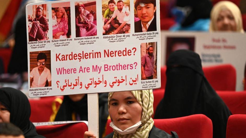 A Muslim Uyghur woman holds up a sign with pictures of her brother and the word: Where are my brothers?