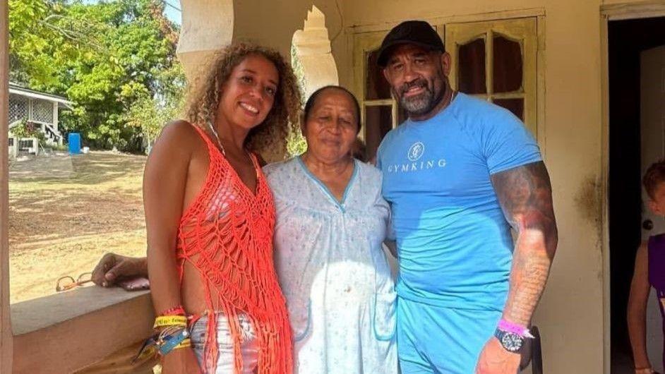 A man wearing a light blue top has his arm around a woman with dark hair in a ponytail and white dress. Next to her is a woman with curly blonde and brown hair and a red mesh top. They are standing outside in a porch with greenery in the distance.