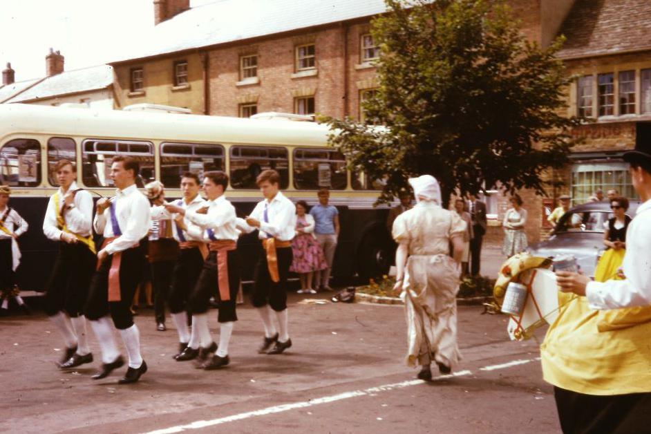 Morris dancers in traditional outfits in front of an old bus, and also someone in a horse outfit, with people watching in the background.