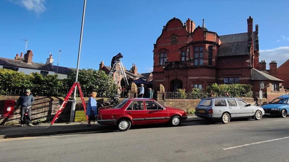 Vintage vehicles are parked along the street outside the old Boys Club on Overleigh Road in Hanbridge. Two ladders, including one with a camera man at the top, can be seen on the road and in the grounds of the club.