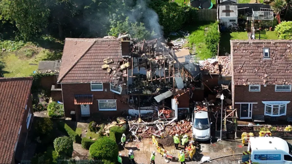 An aerial view of a destroyed semi-detached house surrounded by other houses. Smoke is rising from the building and emergency workers can be seen stood in front of the property.