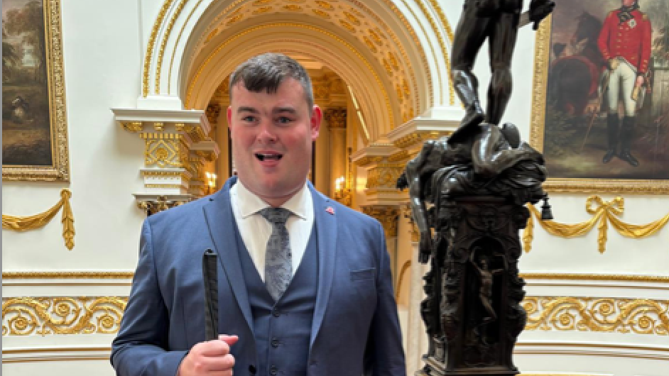 Brandon Hulcoop wears a blue blazer, waistoat and tie with a white shirt. Seen in Buckingham Palace, he smiles holding a stick.