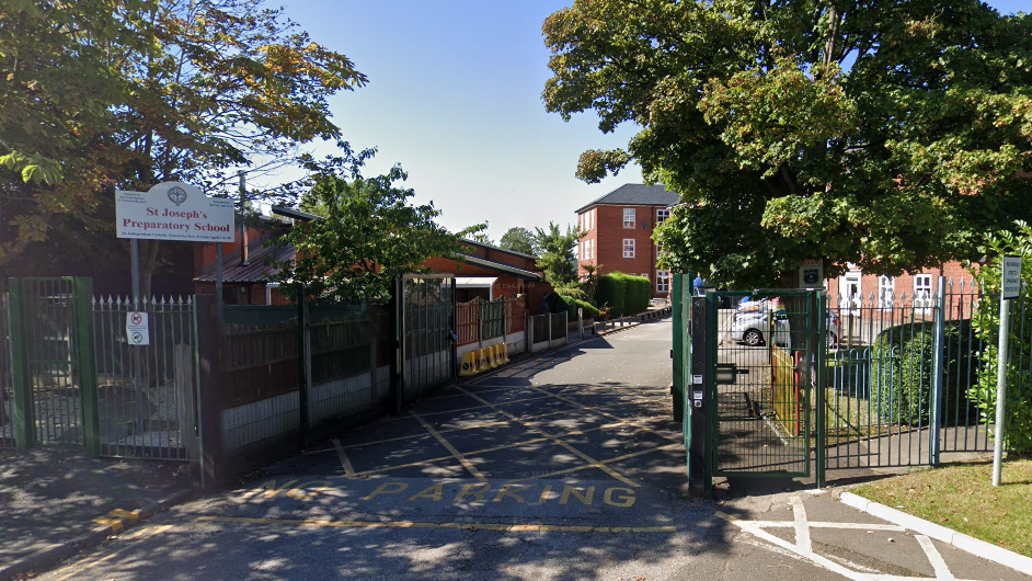 A view of the school gates at St Joseph's Preparatory School. The fencing is in green metal, with a sign at the gate and red brick buildings inside.