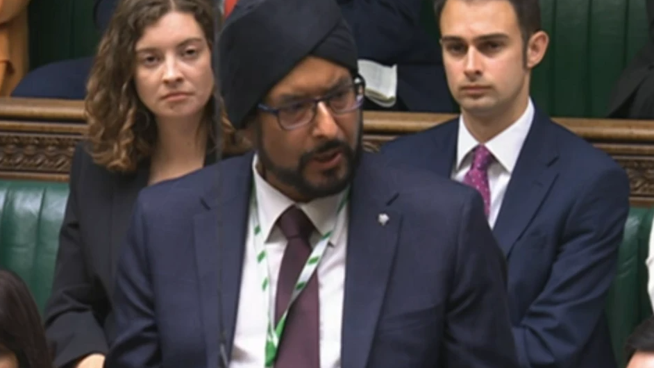 Mr Juss is stood up speaking in Parliament. He wears a turban and glasses and is dressed in a navy suit.