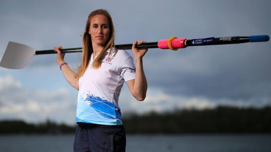 Helen Glover, standing outdoors near a body of water, holding a rowing oar across her shoulders. She has long, light brown hair and is dressed in sportswear, including a white top with blue accents and branding. The background is an overcast sky with distant trees.