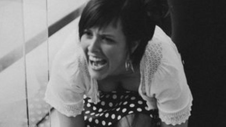 Philippa Reynolds, a woman with short black hair, is crouched down and laughing. She is wearing a white top and spotty skirt.