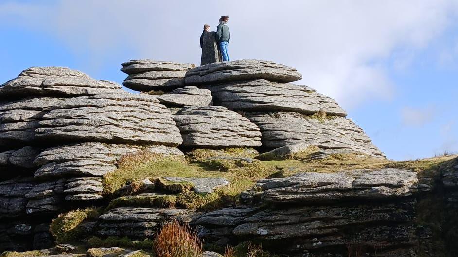 Two people stand on top of a granite tor on Dartmoor. It is a sunny day with light cloud cover. One of the people on the tor appears to be wearing a large cloak.