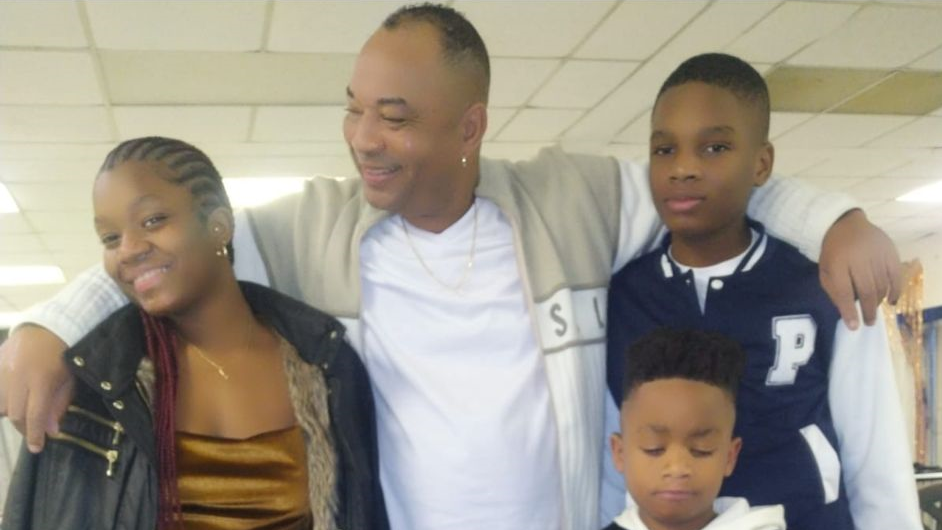 A man posing with his three children who are smiling. The man, in the middle, has a white top and a beige jacket. The teenage children are looking at the camera. The boy on the right is wearing a letterman jacket. The girl on the left is wearing a black jacket. And the smaller boy at the front is looking down. 