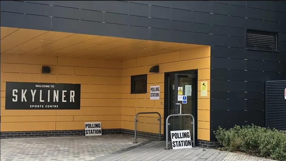 The polling station at Moreton Hall in Bury St Edmunds.  It is a picture of the the outside and front entrance of Skyliner Sports Centre. It has a yellow panelled walls and a black sign. It has a number of "POLLING STATION" signs at the entrance.