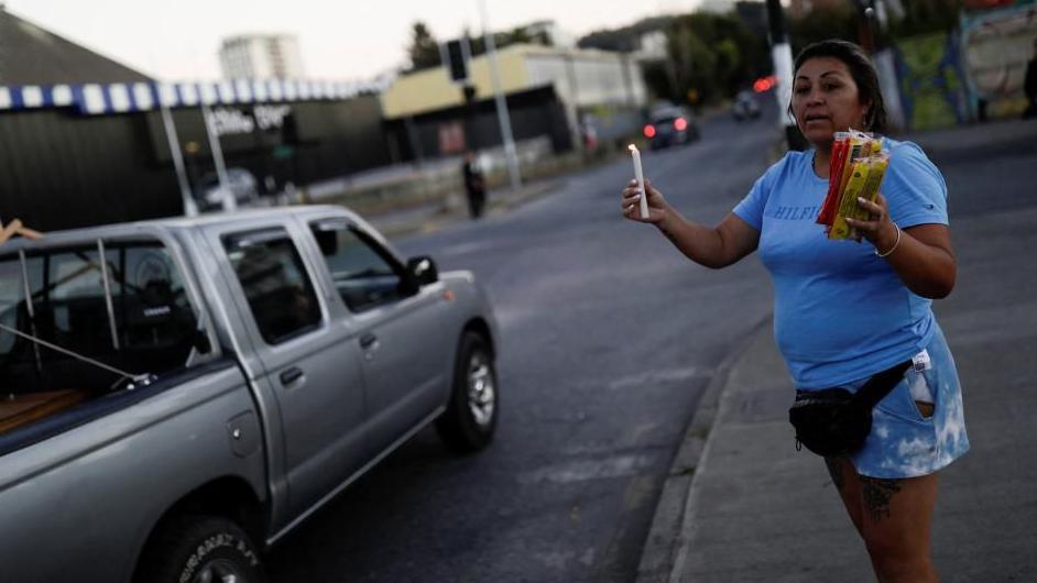 A woman wearing a light blue T-shirt and matching shorts stands on a pavement in Concepción, Chile, holding a lit candle in one hand and packs of candles for sale in the other hand. A silver pick-up truck is cruising past.