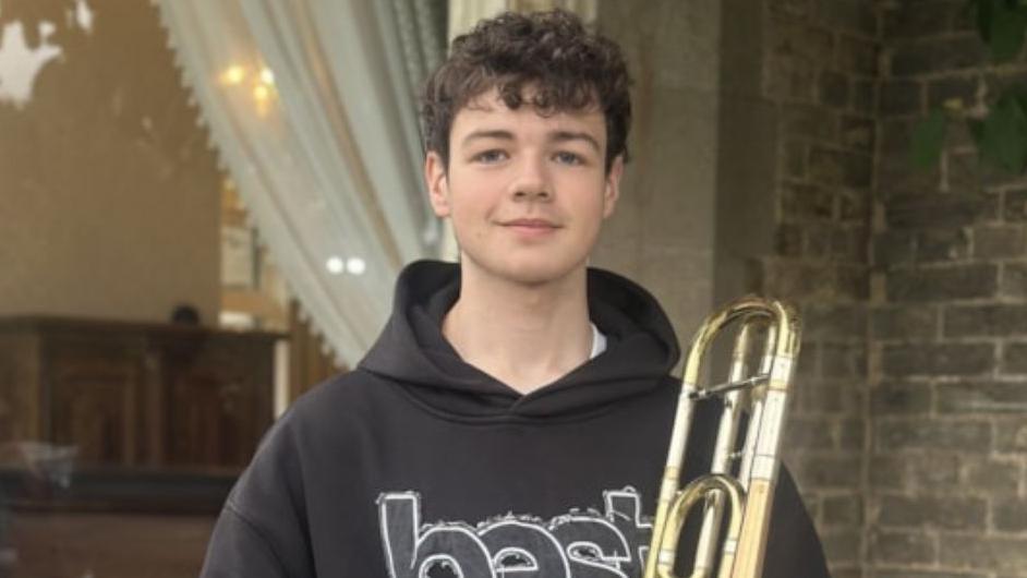 A young man with dark wavy hair, wearing a hooded top and jeans, stands with a trombone. He is smiling. 