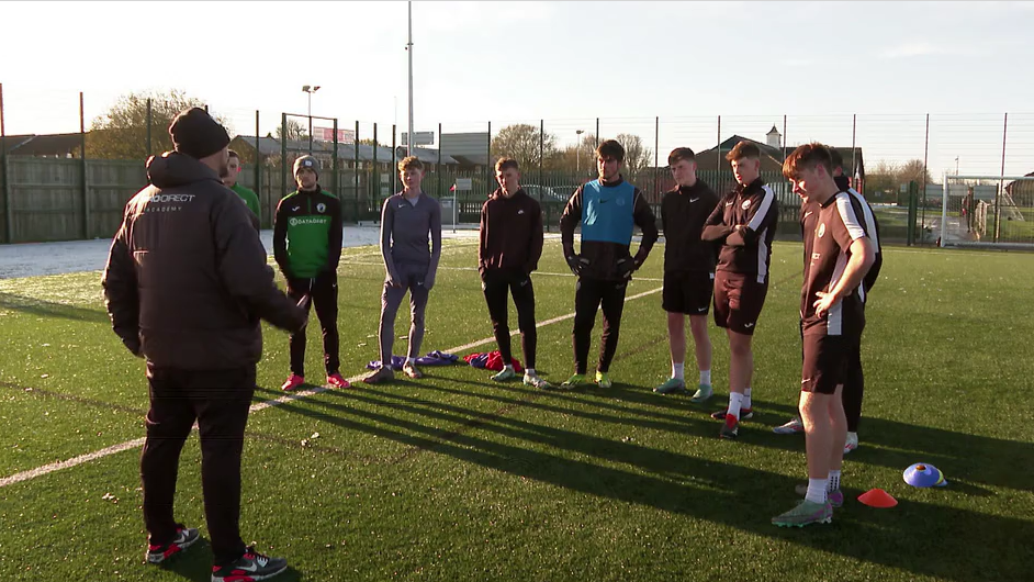 A group of players stand in a circle listening to the coach on a frost-covered pitch. 