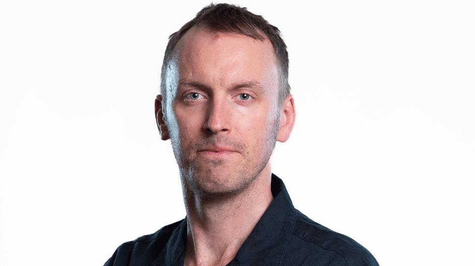 Tom poses for a publicity photo in front of a white background. He wears a black mechanic-style shirt.