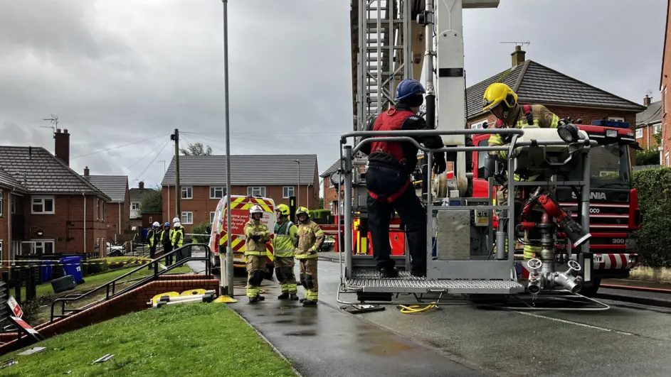 Firefighters stand around at the scene in protective uniform and some in a crane basket near the ground. It is raining and there is is tape cordoning off the scene and a fire engine in the background along with other flats. 