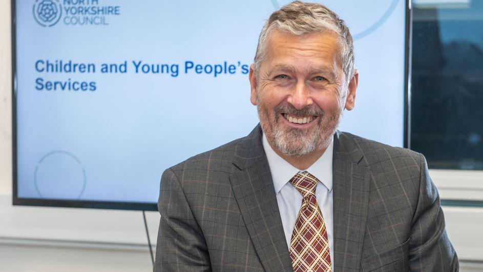 Stuart Carlton, a man with grey hair and wearing a grey suit, shirt and red tie, sits in front a screen which reads 'children's and young people's services'