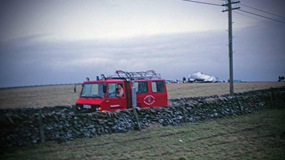 Old faded photo of red rescue vehicle on a road behind a wall in between fields, with the wreckage of a passenger plane in the background.