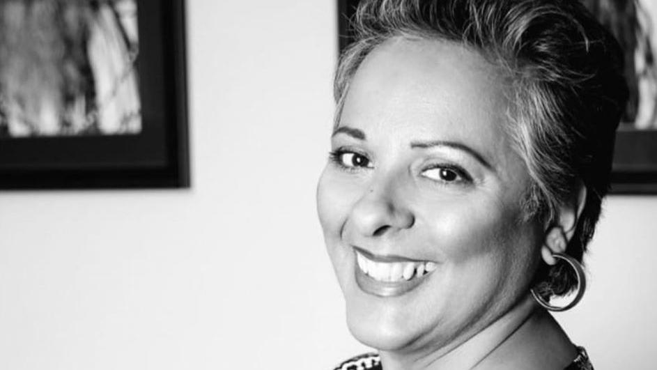 A black and white photo of a woman with dark hair and large hoop earrings. She is smiling at the camera and there are framed pictures on the wall behind her.