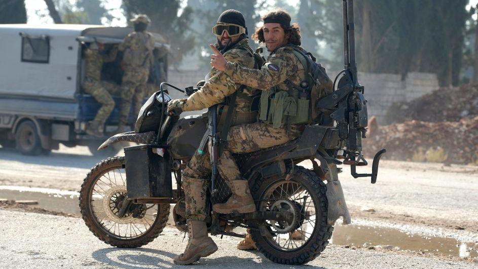 Two men on a scrambler-style motorbike. Both are wearing desert-style camouflage uniforms and ammo belts. The driver has an AK-47 style rifle and there's a large machine gun strapped to the back of the bike. 