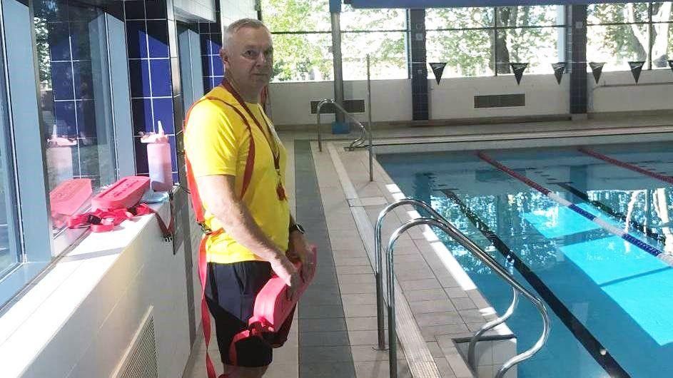 A male lifeguard with grey hair, wearing a yellow t-shirt and carrying a red float, standing by a pool.