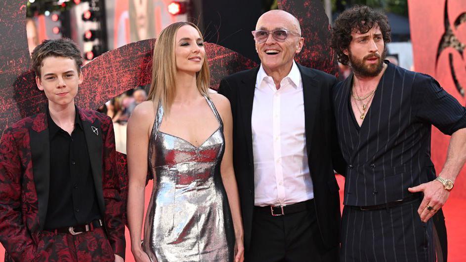 Three actors are standing alongside the director Danny Boyle, Jodie Comer has long blonde hair and is wearing a silver dress, Aaron Taylor-Johnson is in a black suit and has a beard and dark hair. Danny Boyle is in his 60s and is wearing a white shirt